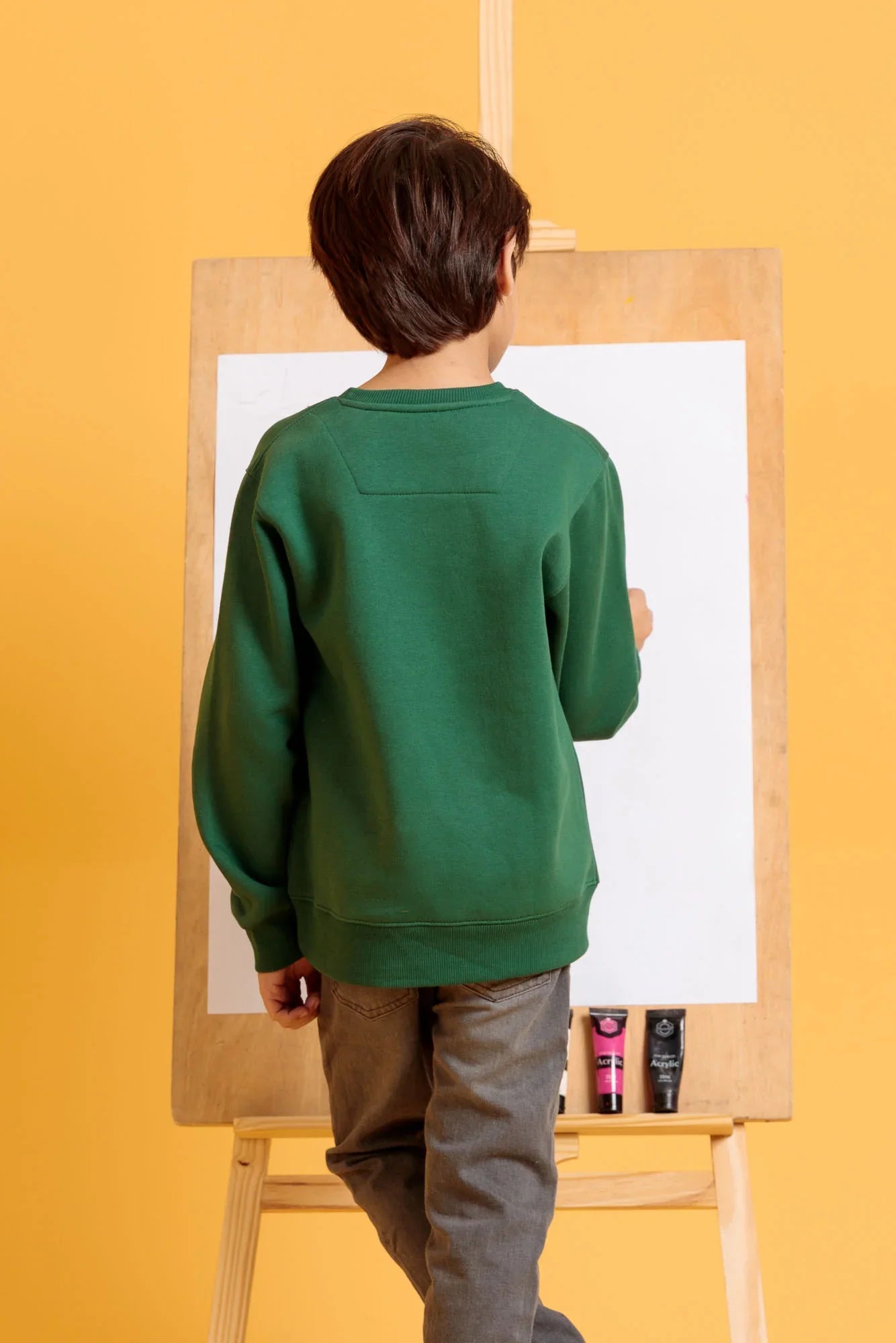 Child in a green sweater standing at an easel with a yellow background