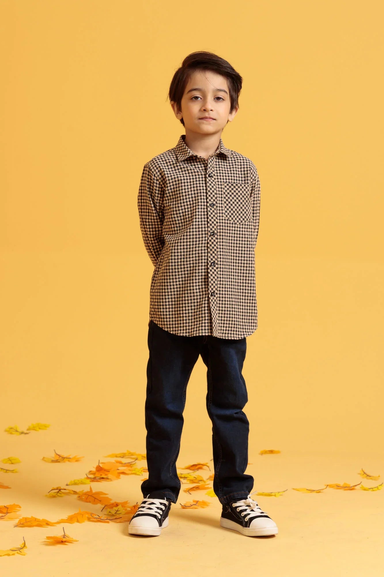 Child wearing a checkered shirt and jeans standing on a yellow background with scattered leaves.