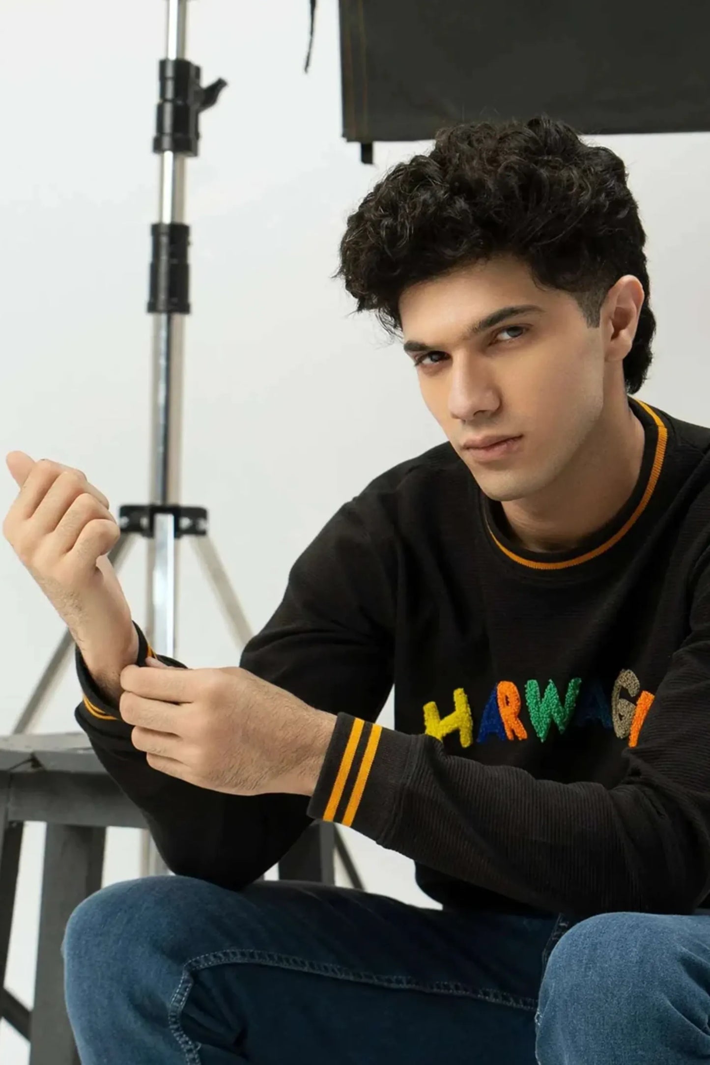Person wearing a black sweatshirt with colorful text, sitting in a studio setting
