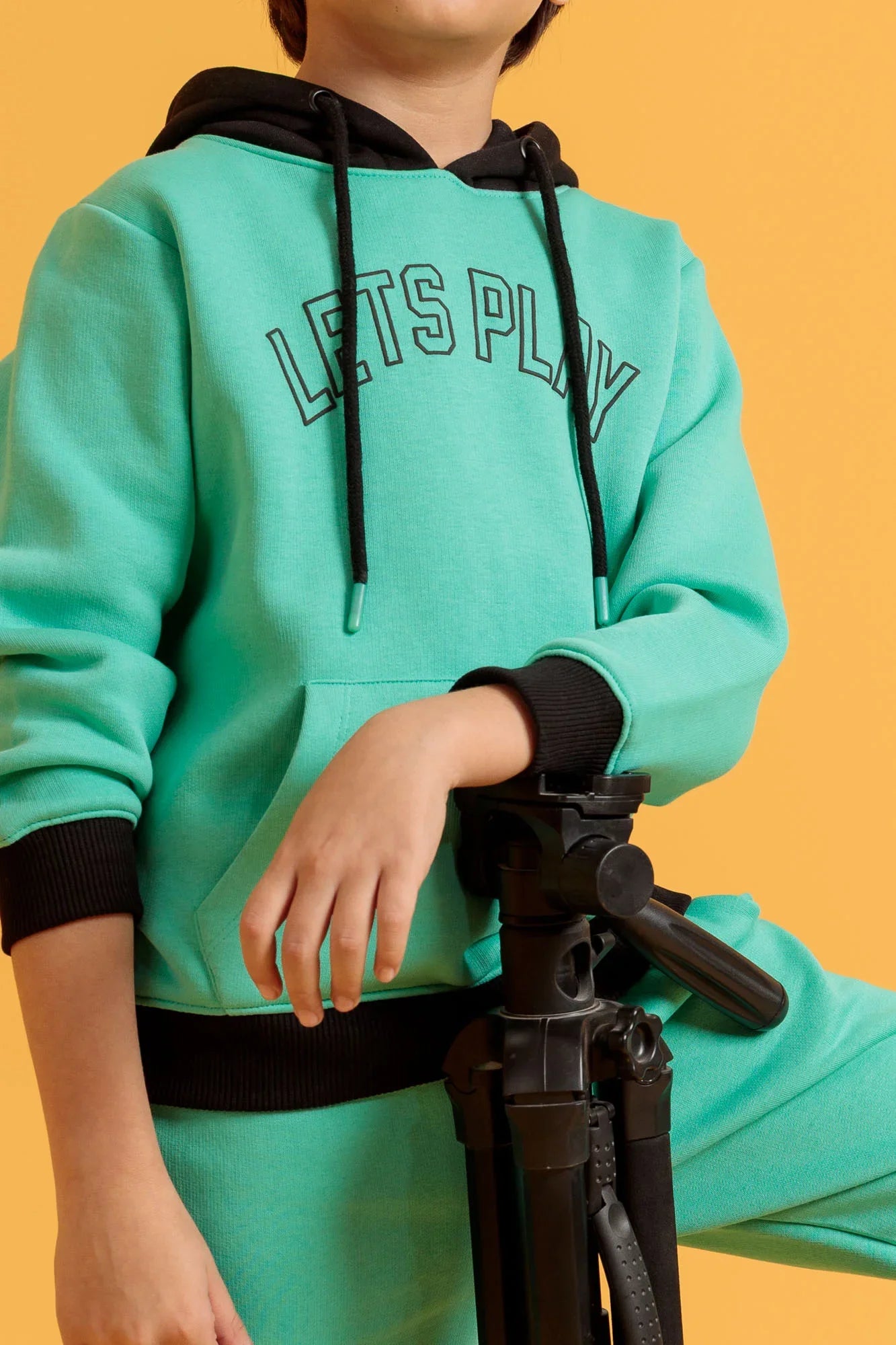 Person wearing a teal hoodie with 'LET'S PLAY' text, sitting on an orange background